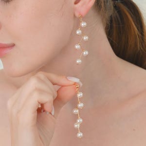 Puede incluir: Pendientes dorados con un diseño en cascada de perlas blancas. Los pendientes presentan una delicada estructura en forma de vid, con múltiples perlas suspendidas a lo largo de la longitud. Los pendientes están diseñados para colgar y crear movimiento.