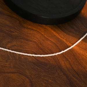 May include: A fine white pearl necklace, set against a warm, wooden backdrop. The necklace features small, uniform pearls, forming a simple, elegant strand. The wooden surface displays a rich, brown hue with visible grain patterns.