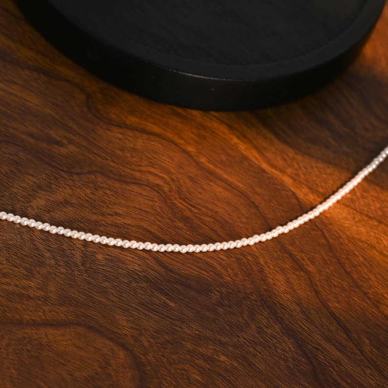 May include: A fine white pearl necklace, set against a warm, wooden backdrop. The necklace features small, uniform pearls, forming a simple, elegant strand. The wooden surface displays a rich, brown hue with visible grain patterns.