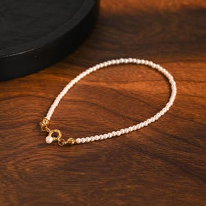 May include: A fine pearl bracelet with a gold clasp, displayed on a dark wood surface. The bracelet is composed of small, round pearls, offering a timeless and refined aesthetic. The gold clasp provides a luxurious accent to the piece.