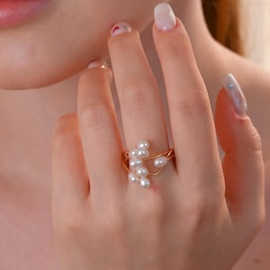 Puede incluir: Un anillo dorado con un grupo de pequeñas perlas blancas. El diseño del anillo incorpora delicados hilos de metal entrelazados que sostienen las perlas. El anillo se lleva en un dedo, mostrando su estética elegante y orgánica.
