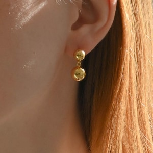 May include: A close-up of a gold-colored earring with two spherical, dangling elements. The earring is worn on an ear, with the person's hair visible in the background. The earring has a shiny, reflective surface.