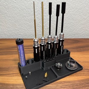May include: A black plastic organizer with six slots for holding screwdrivers. The organizer has a small tray for holding small parts. The screwdrivers are black with silver tips and have the brand name "HobbyPark" printed on them. There is a small tube of solder in the background.