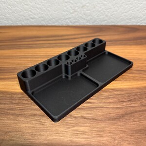 May include: A black plastic organizer with multiple compartments for holding small items. The organizer has a rectangular base with two separate compartments and a vertical section with 10 round holes.