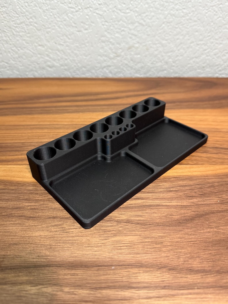 May include: A black plastic organizer with multiple compartments for holding small items. The organizer has a rectangular base with two separate compartments and a vertical section with 10 round holes.