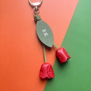 May include: A leather keychain with a gold ring and a green leaf-shaped tag with the initials "S.K." in white. Two red tulip-shaped charms dangle below. The background is orange and green.