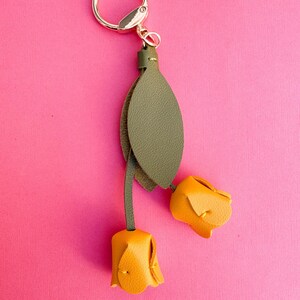 May include: A floral-themed keychain featuring two yellow leather rose buds and a green leather leaf. The keychain has a gold-colored ring and clasp. The background is a solid pink color.