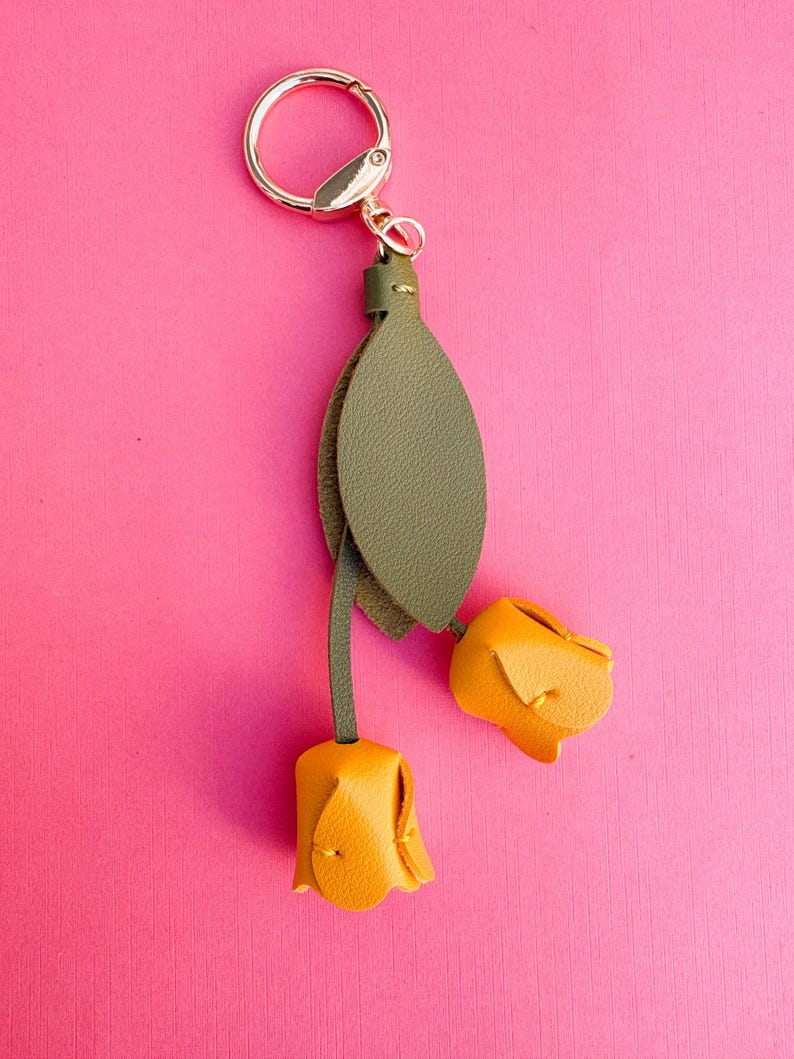 May include: A floral-themed keychain featuring two yellow leather rose buds and a green leather leaf. The keychain has a gold-colored ring and clasp. The background is a solid pink color.