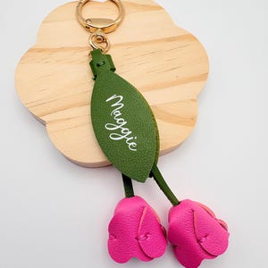 May include: A personalized keychain featuring a green leaf with the name "Maggie" in white script, attached to two pink leather tulip-shaped charms. The keychain has a gold-colored ring for attaching keys.