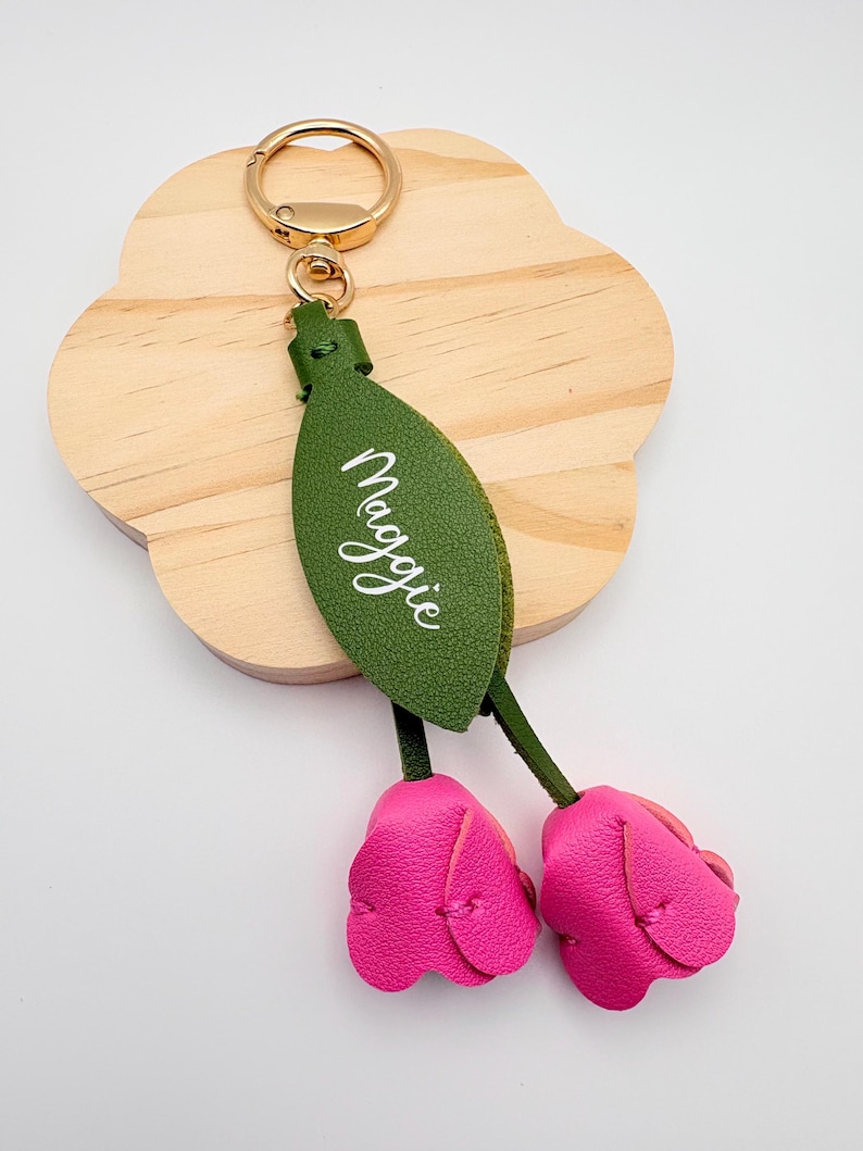 May include: A personalized keychain featuring a green leaf with the name "Maggie" in white script, attached to two pink leather tulip-shaped charms. The keychain has a gold-colored ring for attaching keys.