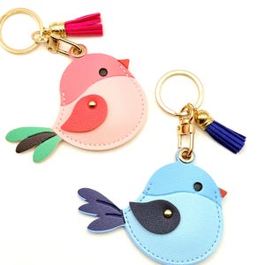 May include: Three bird-shaped keychains in pink, light blue, and blue. Each keychain features a gold-colored ring and clasp, with a tassel in either pink or blue. The birds have contrasting wing and belly colors, with a small black eye and a red beak.