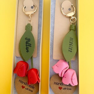 May include: Two keychains in gift boxes. Each keychain features a gold-colored clasp, a green leaf-shaped tag with text, and a leather flower. One has red flowers and the other has pink flowers. Each has a heart-shaped tag that says "Handmade with love."