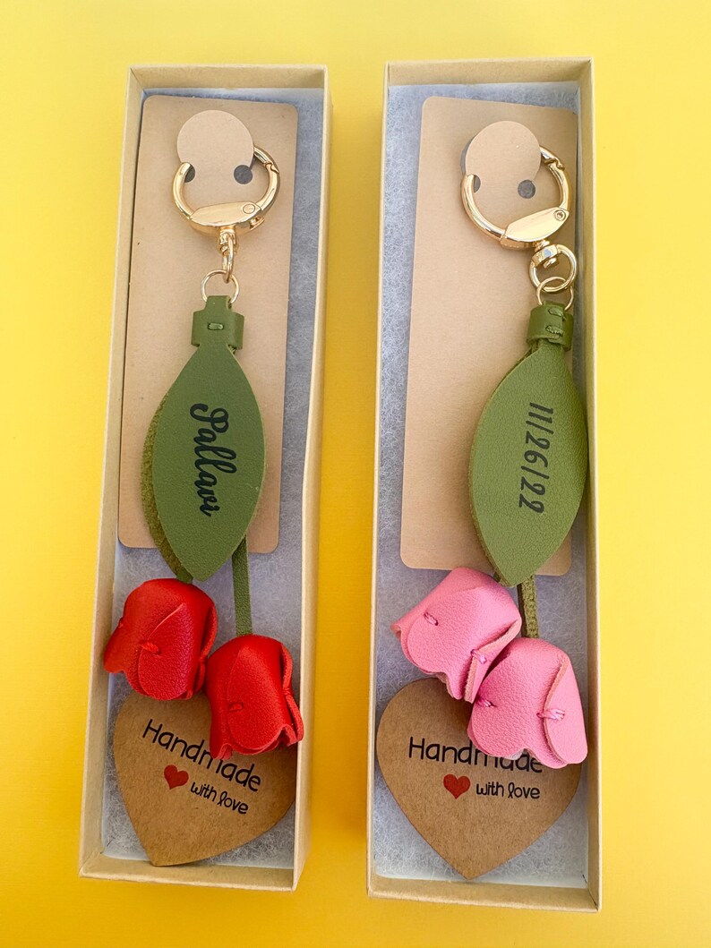 May include: Two keychains in gift boxes. Each keychain features a gold-colored clasp, a green leaf-shaped tag with text, and a leather flower. One has red flowers and the other has pink flowers. Each has a heart-shaped tag that says "Handmade with love."
