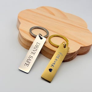 May include: Two rectangular keychains with silver and gold tones. The silver keychain reads "DRIVE SAFE" in black text. The gold keychain has the word "forever" in blue text and a blue heart. Both keychains have a metal ring.