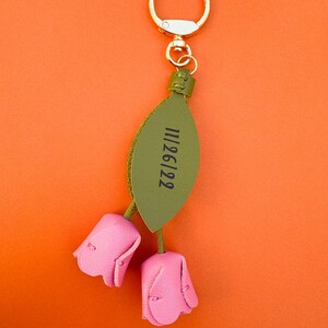 May include: A pink leather keychain with two tulip-shaped flowers and a green leaf. The leaf has the date "11/26/22" printed on it. The keychain has a gold-colored ring for attaching to keys or a bag. The background is orange.