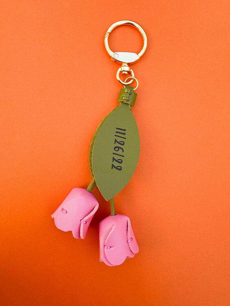 May include: A pink leather keychain with two tulip-shaped flowers and a green leaf. The leaf has the date "11/26/22" printed on it. The keychain has a gold-colored ring for attaching to keys or a bag. The background is orange.