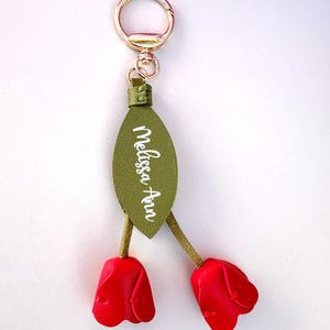 May include: A decorative keychain featuring a gold-colored ring and clasp. The keychain has a green leaf-shaped tag with the name "Melissa Ann" in white script. Two red tulip-shaped charms dangle below.