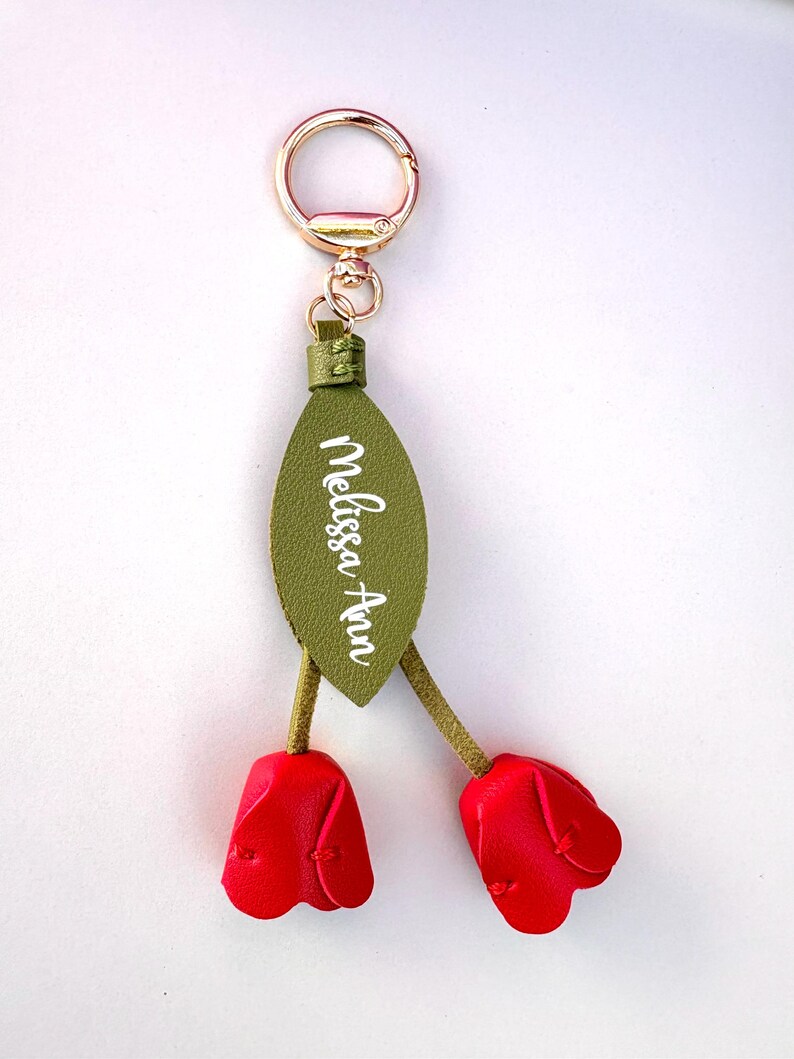 May include: A decorative keychain featuring a gold-colored ring and clasp. The keychain has a green leaf-shaped tag with the name "Melissa Ann" in white script. Two red tulip-shaped charms dangle below.