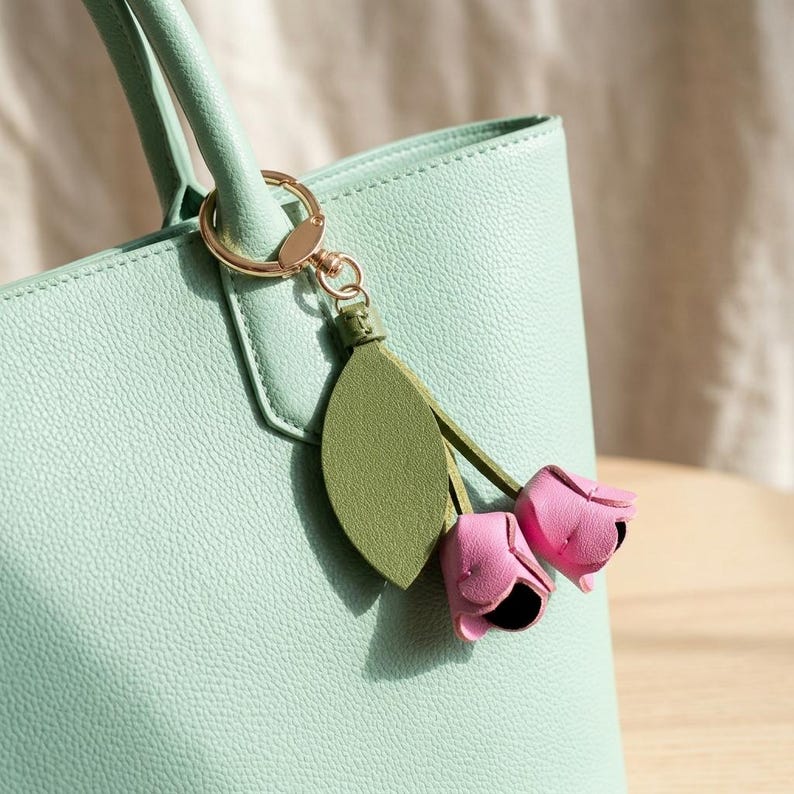 May include: A light green tote bag with a gold-colored keychain and a decorative charm. The charm features a green leaf and two pink flower buds with black centers. The bag has a textured surface.