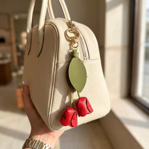 May include: Cream-colored leather handbag with a gold zipper and handles. A decorative charm hangs from the zipper, featuring a green leaf and two red rosebud accents. The bag is held by a person.