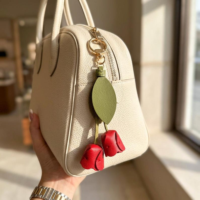 May include: Cream-colored leather handbag with a gold zipper and handles. A decorative charm hangs from the zipper, featuring a green leaf and two red rosebud accents. The bag is held by a person.