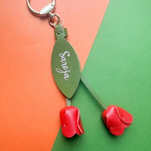 May include: A green leather keychain with the name "Saroja" in white script. The keychain features two red leather tulip-shaped charms and a silver metal ring.
