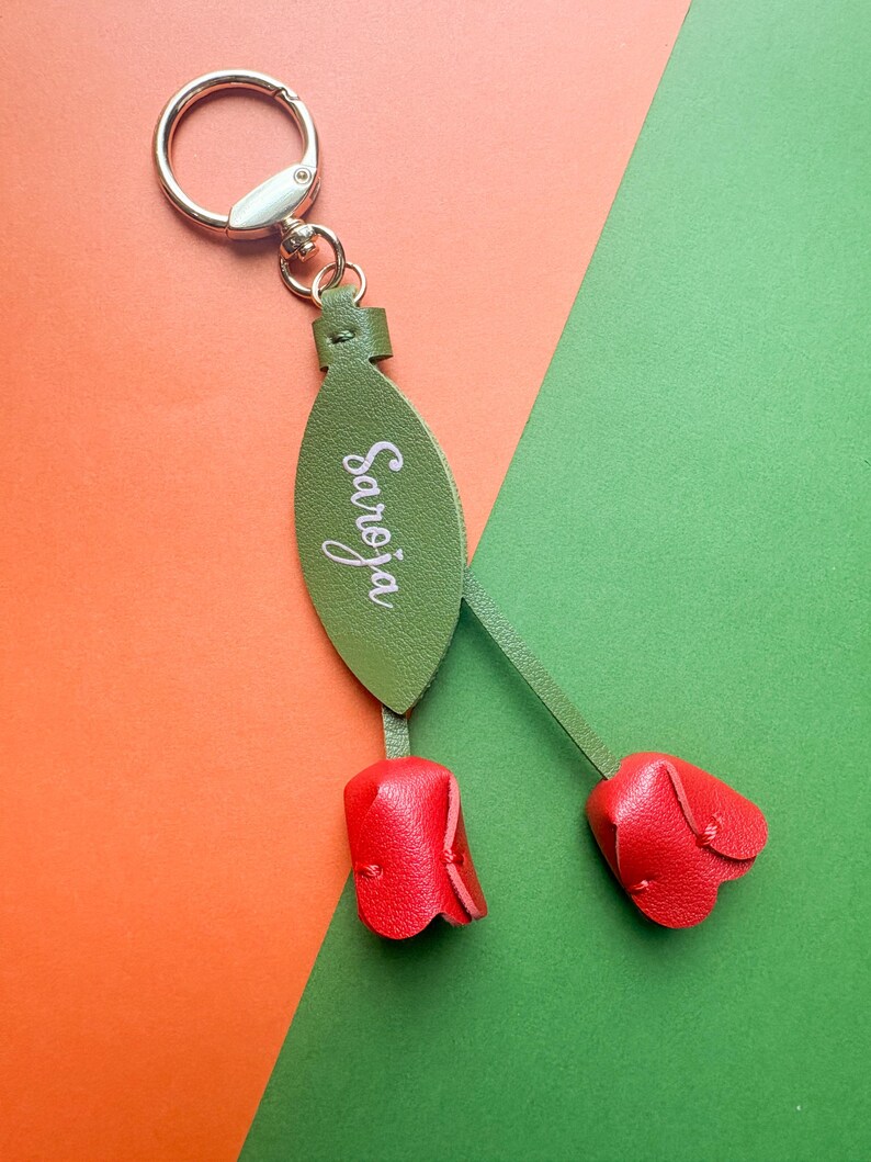 May include: A green leather keychain with the name "Saroja" in white script. The keychain features two red leather tulip-shaped charms and a silver metal ring.