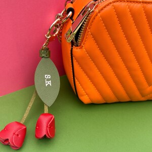 May include: An orange quilted crossbody bag with a gold zipper and chain. A green leaf-shaped charm with the initials "S.K" and two red flower charms hang from the zipper. The bag is on a green and pink background.