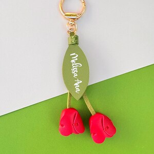 May include: A gold-colored keychain with a green leaf-shaped tag and two red tulip-shaped charms. The tag has the name "Melissa Ann" in white script. The background is white and green.