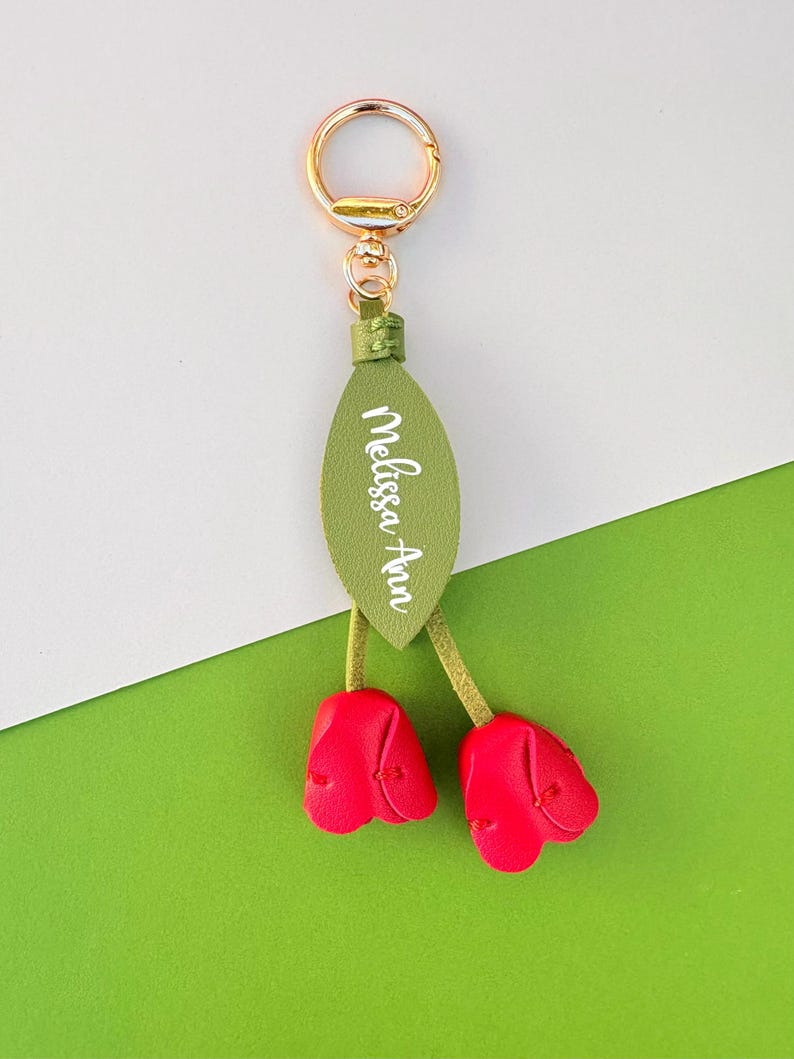May include: A gold-colored keychain with a green leaf-shaped tag and two red tulip-shaped charms. The tag has the name "Melissa Ann" in white script. The background is white and green.