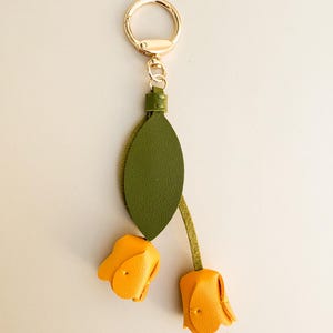 May include: A gold-tone keychain with a green leaf and two yellow tulip flower charms. The keychain has a gold ring and a small green leather accent. The tulip flowers are made of yellow leather.
