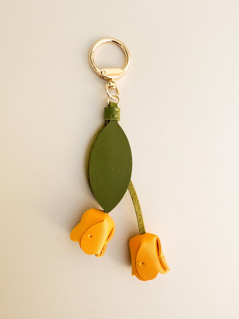 May include: A gold-tone keychain with a green leaf and two yellow tulip flower charms. The keychain has a gold ring and a small green leather accent. The tulip flowers are made of yellow leather.