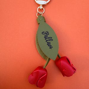 May include: A keychain with a gold-colored ring and clasp. The keychain features a green leaf-shaped tag with the word "Pallavi" in black script. Two red leather rose buds dangle below.