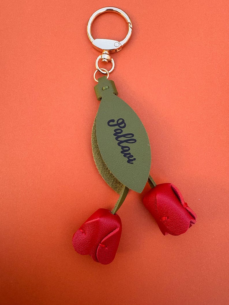 May include: A keychain with a gold-colored ring and clasp. The keychain features a green leaf-shaped tag with the word "Pallavi" in black script. Two red leather rose buds dangle below.