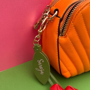 May include: Close-up of an orange, quilted crossbody bag with a gold zipper and hardware. A green leaf-shaped charm with the name "Saroja" in cursive hangs from the bag, along with two small red tulip-shaped charms. The background is pink and green.