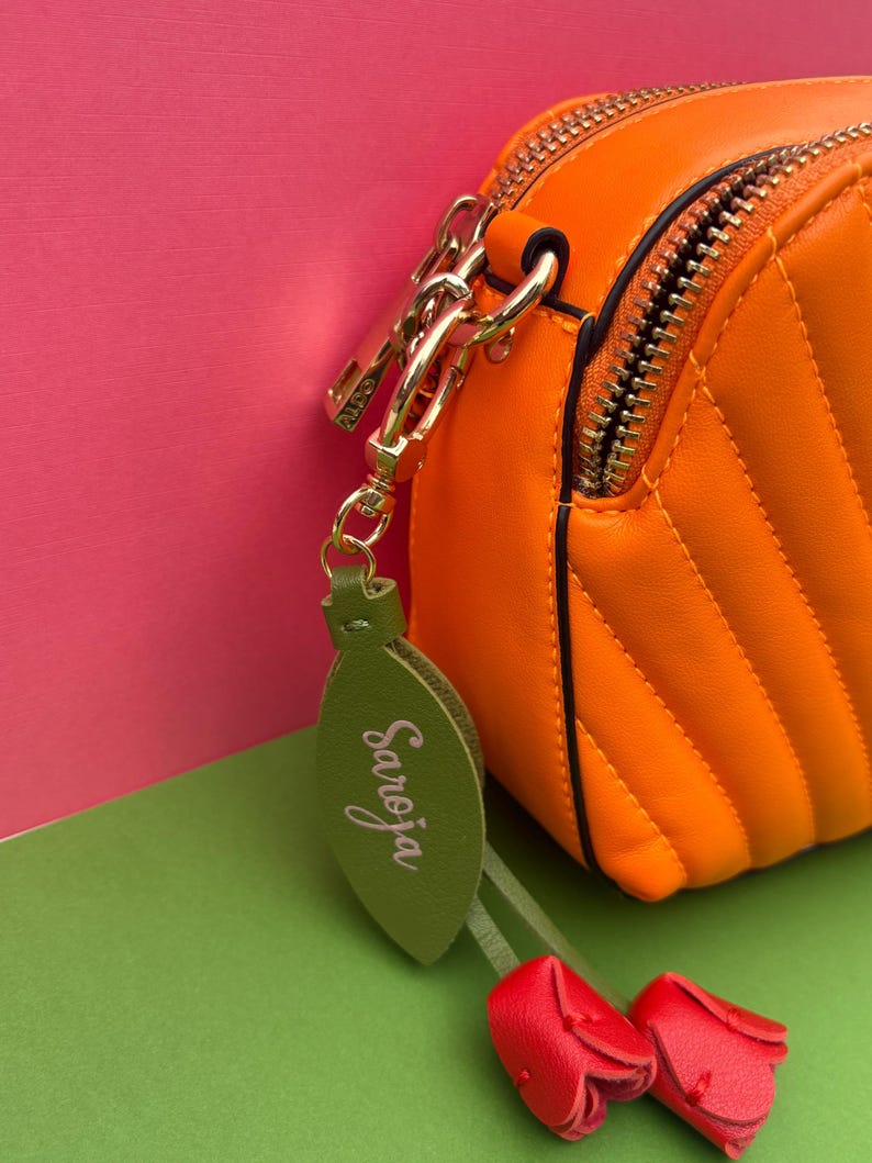 May include: Close-up of an orange, quilted crossbody bag with a gold zipper and hardware. A green leaf-shaped charm with the name "Saroja" in cursive hangs from the bag, along with two small red tulip-shaped charms. The background is pink and green.