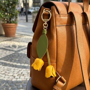 May include: A brown leather backpack with a gold-colored clasp and straps. A decorative charm with yellow tulip flowers and a green leaf hangs from the clasp. The charm is attached with a gold-colored chain.