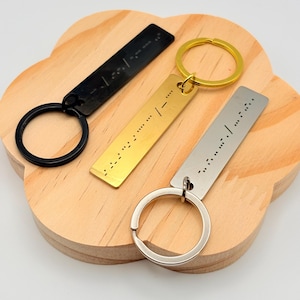 May include: Three metal keychains in black, gold, and silver, each with a rectangular tag and a circular keyring. The tags feature a series of raised dots, likely representing Braille. The keychains rest on a light brown, wooden, cloud-shaped tray.