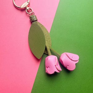 May include: A leather keychain featuring two pink tulip-shaped charms and green leaves. The keychain has a gold-colored ring and clasp. The background is divided diagonally, with pink and green sections.