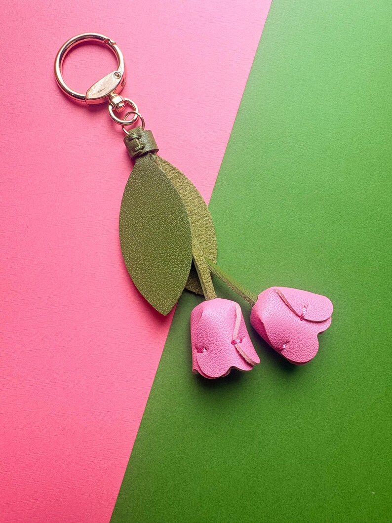 May include: A leather keychain featuring two pink tulip-shaped charms and green leaves. The keychain has a gold-colored ring and clasp. The background is divided diagonally, with pink and green sections.
