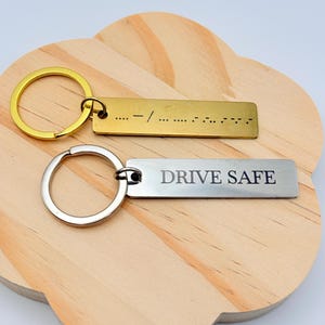 May include: Two rectangular keychains. One is gold-colored with a gold ring and Morse code. The other is silver-colored with a silver ring and the words "DRIVE SAFE". Both keychains are on a wooden surface.