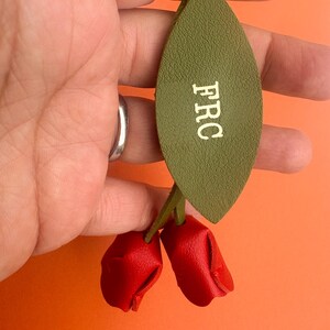 May include: Olive green leather keychain with the letters "FRC" in white. The keychain features two red leather rose buds. The background is orange.