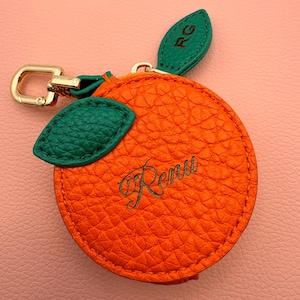 May include: Orange-shaped keychain with green leaf accents and a gold-colored clasp. The orange-colored leather has a textured surface and the name "Renu" is written in cursive. The green leaves have the initials "RG".