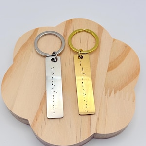 May include: Two rectangular keychains with Morse code. One is silver with a silver ring, the other is gold with a gold ring. Both keychains are on a wooden, cloud-shaped tray. The keychains have a series of dots and dashes.