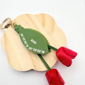 May include: A leather keychain featuring two red tulip blossoms and a green leaf-shaped tag. The tag has the text "2026" and coordinates. The keychain has a gold-colored ring.