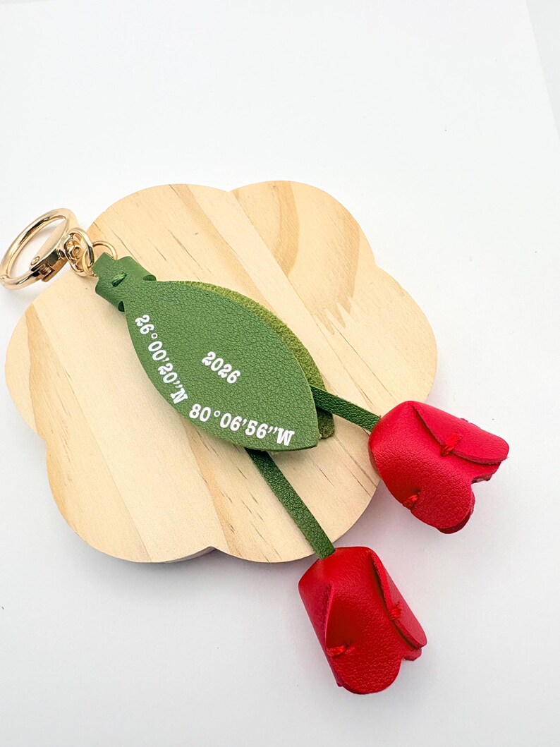 May include: A leather keychain featuring two red tulip blossoms and a green leaf-shaped tag. The tag has the text "2026" and coordinates. The keychain has a gold-colored ring.