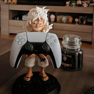 Puede incluir: Figura de un personaje de manga con pelo blanco y rostro sonriente, sosteniendo un mando de PlayStation 5 blanco y negro. La figura está sobre una base negra. A la derecha hay un tarro de vela negro con tapa de cristal.