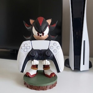 May include: A Shadow the Hedgehog figurine holding a white and black PlayStation 5 controller. The figurine has red and black accents and is standing on a green and brown base. A PlayStation 5 console is visible in the background.