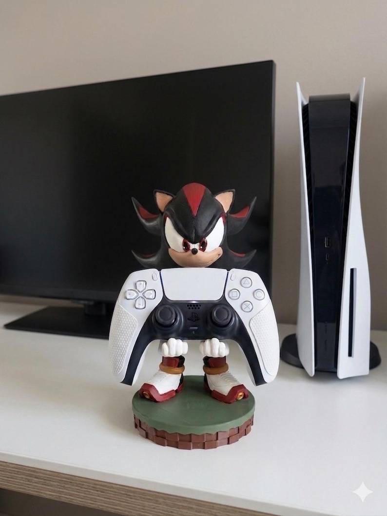 Peut inclure: Figurine de Shadow the Hedgehog tenant une manette PlayStation 5 blanche et noire. La figurine a des accents rouges et noirs et repose sur une base verte et marron. Une console PlayStation 5 est visible en arri&egrave;re-plan.