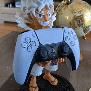 May include: A figurine of a character with spiky white hair and a wide grin, standing on a black circular base with the text GEAR 5. The character is holding a white and black PlayStation 5 game controller in their hands, positioned in front.